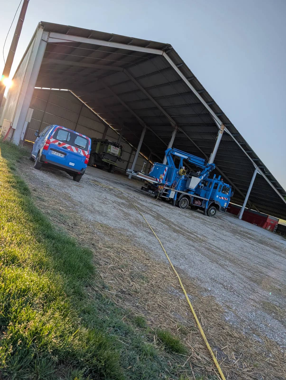 Hangar agricole ouvert, un fourgon bleu et un camion nacelle bleu garés dessous, un tuyau jaune sur le gravier